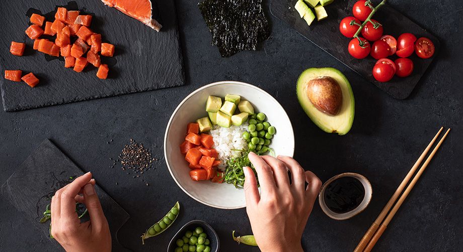 Mani che preparano Poké Bowl su ciotola bianca con riso, salmone e verdure spezzettate